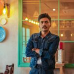 Young man with mustache in a denim jacket posing indoors with reflective window and wall decor.